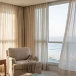 Fotografia realista de uma sala de estar luxuosa com vista para o mar, destacando cortinas claras de tecido nobre filtrando a luz do sol. A imagem ilustra a constante exposição das fibras naturais ao clima litorâneo, reforçando a importância da lavagem de cortinas em Itapema para proteger a decoração de alto padrão contra a oxidação, os raios UV e os danos causados pela maresia.