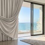Fotografia realista de uma sala de estar moderna com vista para o mar em Itapema, destacando uma cortina blackout branca com forro térmico que protege o ambiente do sol forte do litoral.